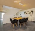 Modern dining area with a wooden table, black chairs, and a potted plant. The room has a white wall, a ceiling with lights, and a refrigerator on the right side.