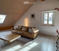 Modern living room with a brown leather sofa, wooden coffee table, and dining area with chairs and table. The room features wooden slanted ceilings, large windows, and a skylight.