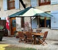 Outdoor patio area with a wooden table, six chairs, and a large umbrella, set against a stone house with shutters and a flag.
