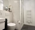Modern bathroom with white tiles, a toilet, sink, mirror, shower, radiator, and a decorative flower vase on the sink counter.
