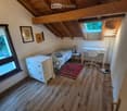 Cozy attic bedroom with wooden slanted ceilings, a single bed, a nightstand with a lamp, a small desk with a chair, a drawer unit, and a floor lamp. The room features wooden flooring and large windows with a view.
