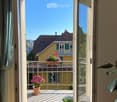 Open balcony with wooden floor, metal railing, and potted plants, overlooking a clear blue sky and neighboring houses.