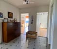 A clean, well-lit hallway with tiled flooring and white walls. Features include a wooden cabinet with framed pictures, a small wooden pet bed, and several doors leading to other rooms.