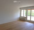 Empty room with light wooden flooring, white walls, large windows, and a radiator under one window.
