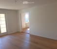 Empty room with light wooden flooring, white walls, and large windows with white frames. The room has a sliding glass door leading to a balcony or outdoor area.