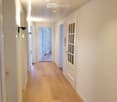 Bright hallway with light wooden flooring, white walls, and several doors leading to other rooms. The ceiling features recessed lighting and a white panel design.