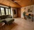 A cozy living room with a green sectional sofa, wooden coffee table, laptop on a desk, floor lamp, and several potted plants. The room features large windows with a view of greenery outside, a wooden door, and a warm, inviting atmosphere.