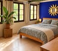 Cozy bedroom with a wooden bed, nightstand, lamp, and potted plants. Blue accent wall with a sun-shaped mirror.