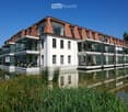 Modern two-story building with white walls, red-tiled roof, and large glass balconies overlooking a serene water body.