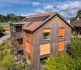 Modern two-story house with wooden exterior, solar panels on the roof, and a balcony with outdoor furniture.