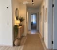 Modern hallway with wooden flooring, white walls, and soft lighting. Features a small wooden console table with a lamp, decorative plants, and a woven rug leading to an open doorway.