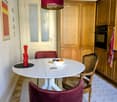 A cozy kitchen with a round white dining table, red chairs, a wicker chair, a bowl of fruit, a pepper grinder, wooden cabinets, and an oven.