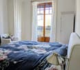 Cozy bedroom with a large bed covered in a floral-patterned duvet, a nightstand with a lamp, a wooden door leading to a balcony, and a small armchair with a patterned pillow.
