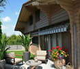 A well-maintained patio area with two armchairs, a small round table, and a vase of colorful flowers. The patio is adjacent to a house with a striped awning and is surrounded by lush greenery and potted plants.