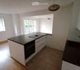 Modern minimalist kitchen with island, black granite countertop, white cabinets, stainless steel oven, integrated sink, natural wooden flooring, two windows, balcony with railings
