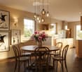 Open-plan dining and kitchen area with a round wooden table surrounded by six wooden chairs, a vase of pink and white flowers, framed artwork on the walls, and a modern pendant light fixture above the table. The kitchen features white cabinets, a sink, an