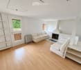 Modern living room with light-colored wooden flooring, white walls, and a fireplace. Features two beige sofas, a white shelf unit, and a small window with a view of greenery.