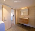 Modern bathroom with light wooden cabinet, sink, and mirror. The room features light-colored tiles and a wooden door leading to another space.