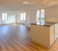 Empty kitchen with light wooden flooring, white walls, and a modern kitchen island with a sink and faucet. Large windows provide natural light.