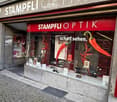 Storefront of an optician shop named "Stampfli Optik" with a red awning, glass display case, and various optical products visible inside.
