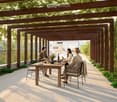 Outdoor dining area with wooden table, chairs, and a pergola covered with greenery, overlooking a scenic valley.