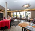 Restaurant interior with multiple tables and chairs, wine glasses, cutlery, and napkins set on tables, large windows with outdoor view, and a refrigerator in the background.
