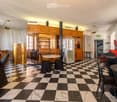 A spacious, elegantly furnished room with a black and white checkered marble floor, multiple tables and chairs, a refrigerator, wooden cabinets, and large windows with curtains.