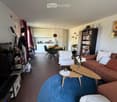 Modern living room with a blue round rug, a brown sofa, a white armchair, wooden coffee tables, a bookshelf, and a dining area with a round table and chairs. The room features white walls, wooden flooring, and a kitchen area with white cabinets.