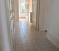 A clean, tiled hallway with white walls, multiple doors, and a slanted wooden ceiling. Natural light streams in from an open doorway at the end.