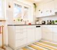 Bright, modern kitchen with white cabinets, tiled walls, and a dark countertop. Features a dishwasher, faucet, potted plants, and decorative items on the counter.