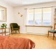 Bright bedroom with wooden flooring, large window with white curtains, green armchair, small wooden table with plant, and a mirror on the wall.