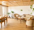 Bright living room with wooden flooring, a large sofa, armchairs, a dining table with chairs, a bookshelf, and potted plants.