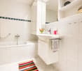 Modern bathroom with white tiles, a bathtub, a sink with a mirror, a colorful striped rug, and built-in cabinets.