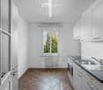 Modern kitchen with white cabinets, stainless steel sink, oven, and tiled floor. Large windows provide natural light.
