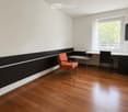 A modern, minimalist room with wooden flooring, white walls, and a large window with white curtains. Features include a black and white accent wall, a red chair, a small table, and a mounted screen.