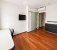 A modern, minimalist room with wooden flooring, a white table, a single chair, and a wall-mounted TV. Features a sliding glass door leading to another room.
