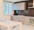 Modern kitchen with light wood cabinets, stainless steel appliances, white countertops, and a dining area with a white table and transparent chairs.