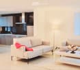 Modern open-plan living and dining area with white walls, light wood flooring, and large windows. Features a white sofa with a red throw, a coffee table with books and a decorative bowl, a dining table with chairs, and a sleek kitchen area with cabinets a