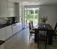 Open-plan kitchen and dining area with white cabinets, a dark dining table with chairs, and a vase of flowers on the table. The kitchen features a sink, oven, and dishwasher, with a view to a garden through large glass doors.
