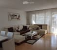 Modern living room with a sectional sofa, coffee table, dining chairs, and artwork on the wall. The room features large windows with sheer curtains and a wooden floor.