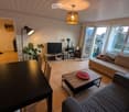 Bright living room with large windows offering a view, featuring a sofa, coffee table, TV, dining table, chairs, floor lamp, and a plant.