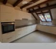 Modern kitchen with white cabinets, stainless steel oven, and a sink with a faucet. The ceiling features exposed wooden beams and skylights.