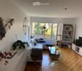 Modern living room with a white sectional sofa, glass coffee table, bookshelf, cat tree, and decorative plants. Large windows provide natural light.
