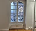 Empty room with wooden flooring, white walls, a large arched window with decorative glass, and a radiator below the window.