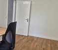 A small, unfurnished room with a wooden desk and a black office chair, white walls, and a closed white door.