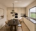 Modern kitchen with light-colored cabinets, marble countertops, a central dining table with chairs, and a pendant light fixture. Large windows offer a scenic view of the countryside.