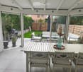 A covered patio with a dining table, chairs, and a vase with dried flowers. The space has large glass doors opening to a garden with a sofa, potted plants, and a barbecue.