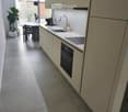 Modern kitchen with white cabinets, stainless steel sink, built-in oven, and a small dining area with a table and chairs. The space features tiled flooring and large windows with blinds.