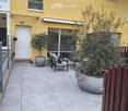 A small outdoor patio area with a tiled floor, a white door leading to the building, two potted olive trees, a lounge chair, and a small table.