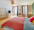 Bright bedroom with wooden floor, large windows, balcony access, bed with colorful blankets, dresser, chair, and screen.